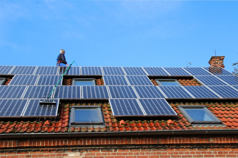 Bei der Solaranlagenreinigung: Gebäudereiniger oben auf dem Dach, hält den Stiel der Reinigungsbürste; Solarpaneele auf dem Dach.